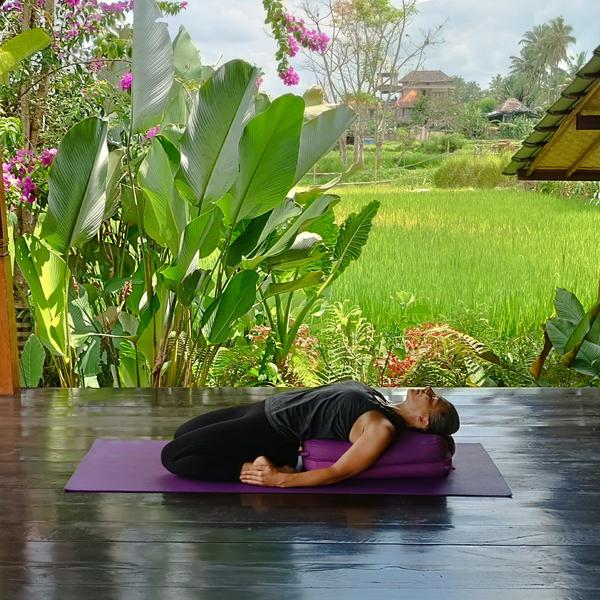 Eine Person übt eine erholsame Yoga-Pose aus und liegt auf einer lila Yogamatte mit einem Polster unter dem Kopf und dem oberen Rücken, die Arme an den Seiten, in einer grünen Umgebung mit üppiger Vegetation und einem Feld im Hintergrund.