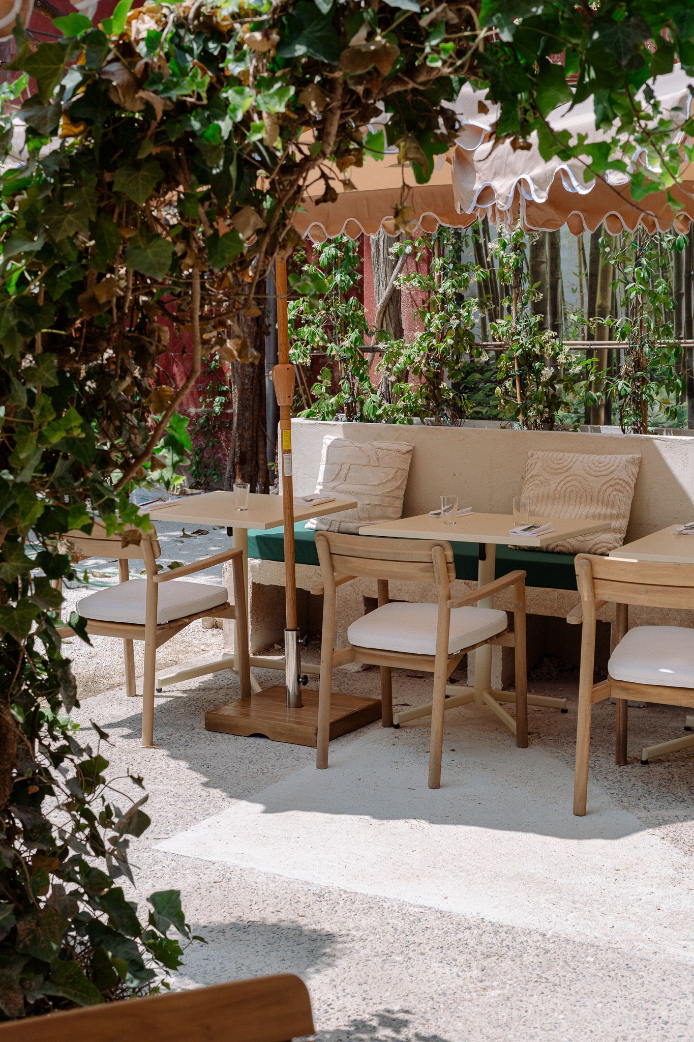 Café-Terrasse im Schatten von Grün und Sonnenschirm mit hellen Holztischen und gepolsterten Sitzen.