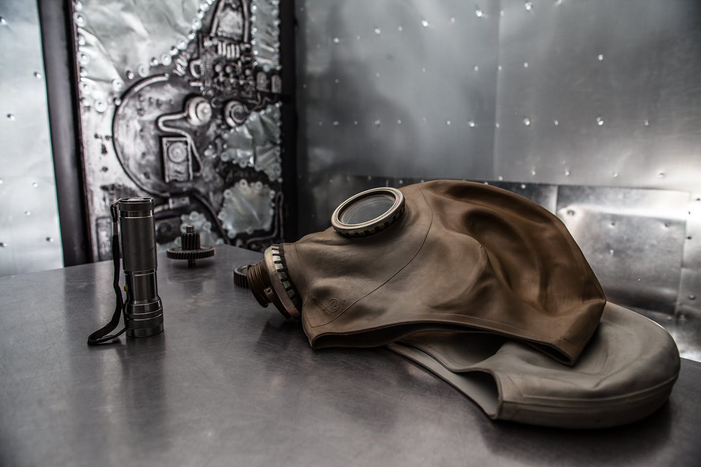 A vintage beige gas mask and a metal flashlight are placed on a metal table , in front of a textured industrial or sci-fi wall . 