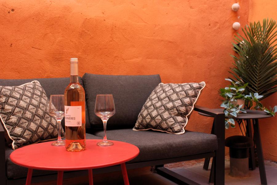 Ein kleiner Innenhof mit orangen Wänden, warm und intim. Ein dunkelgraues Sofa mit zwei schwarzen und weißen geometrischen Kissen nimmt den Hintergrund der Szene ein. Davor stehen auf einem runden roten Couchtisch eine Flasche Roséwein und zwei Stielgläser.. Auf der rechten Seite befindet sich ein Beistelltisch mit einer grünen Topfpflanze, die für einen Hauch von Frische sorgt. Die Atmosphäre erinnert an einen Moment geselliger Entspannung, perfekt für einen Aperitif. 