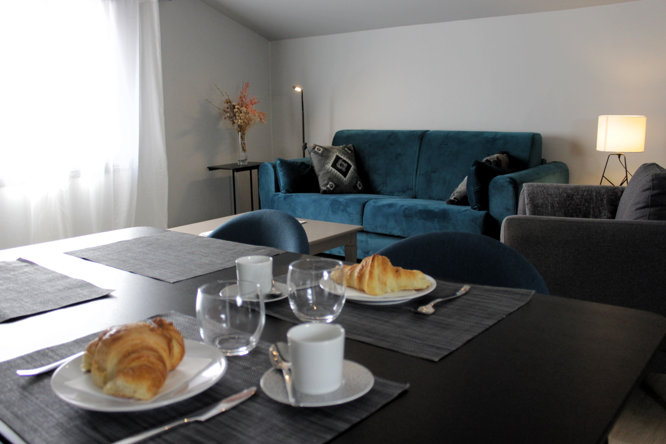 Ein helles, modernes Zimmer, das in einem einfachen und eleganten Stil eingerichtet ist. Im Vordergrund ist ein schwarzer Frühstückstisch aufgestellt. Im Vordergrund ein schwarzer Frühstückstisch mit zwei Tellern mit Croissants, runden Gläsern und weißen Tassen auf grauen Tischsets. Im Hintergrund ist ein türkisfarbenes Samtsofa mit schwarzen und grauen geometrischen Kissen geschmückt. Auf der rechten Seite verbreitet eine Tischlampe weiches Licht, und ein Strauß getrockneter Blumen schmückt einen Couchtisch. Das Set schafft eine einladende und gemütliche Atmosphäre. 