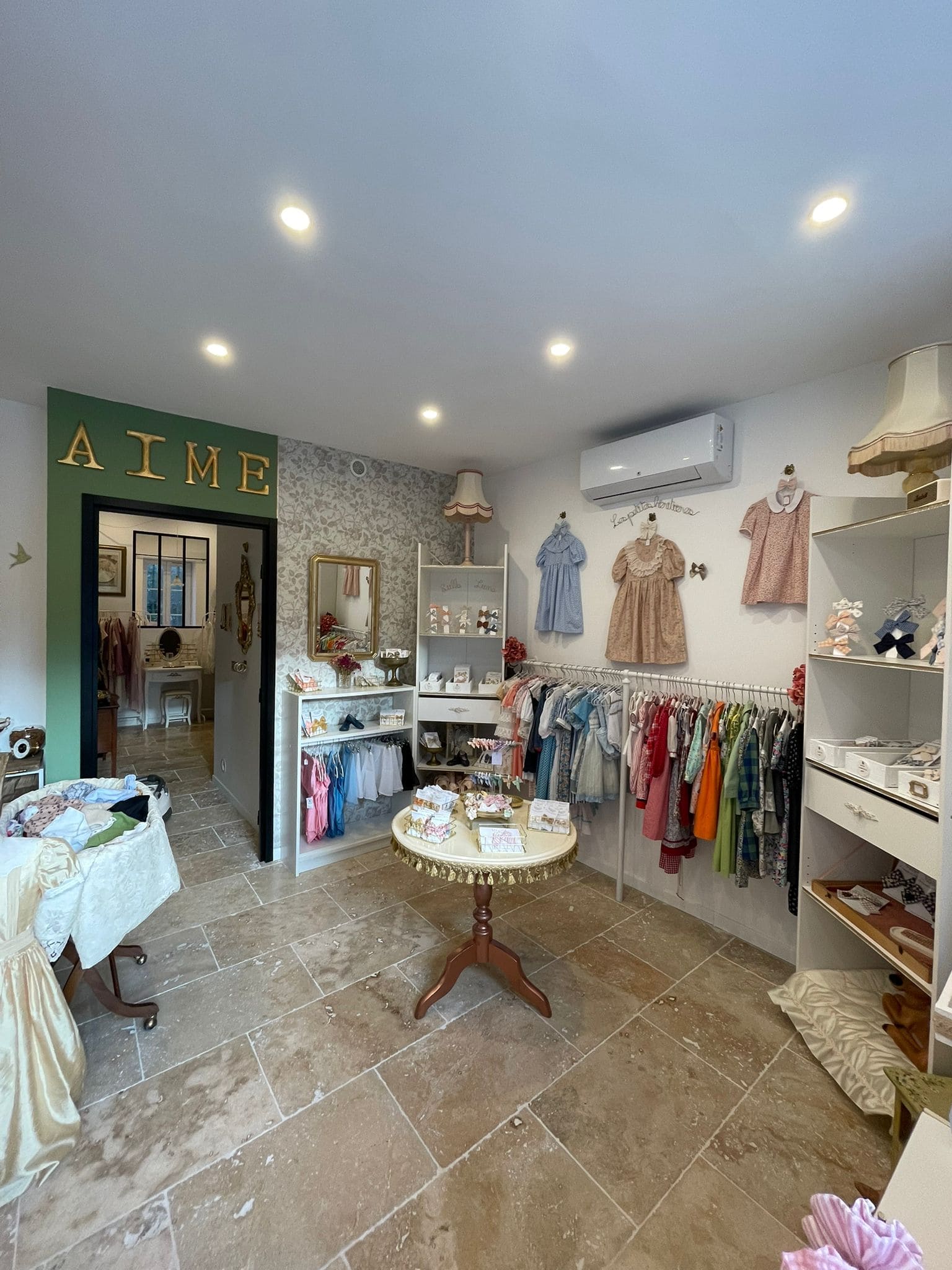 Ein heller Boutique-Raum mit hellem Steinboden und verzierten weißen Wänden. An Kleiderbügeln hängen mehrere Kinderkleider: kleine Kleider, Hemden und farbenfrohe Sets. Auf der rechten Seite zeigt ein Kleiderständer eine Vielzahl von Outfits. Auf der linken Seite ein runder Tisch mit Produkten (wahrscheinlich Accessoires oder kleine Modeartikel). Das Ambiente ist weich und elegant, mit Retro-Details wie einer Lampe mit Stoffschirm und einigen goldenen Titeln, die das Wort LIEBE über der Tür bilden. 
