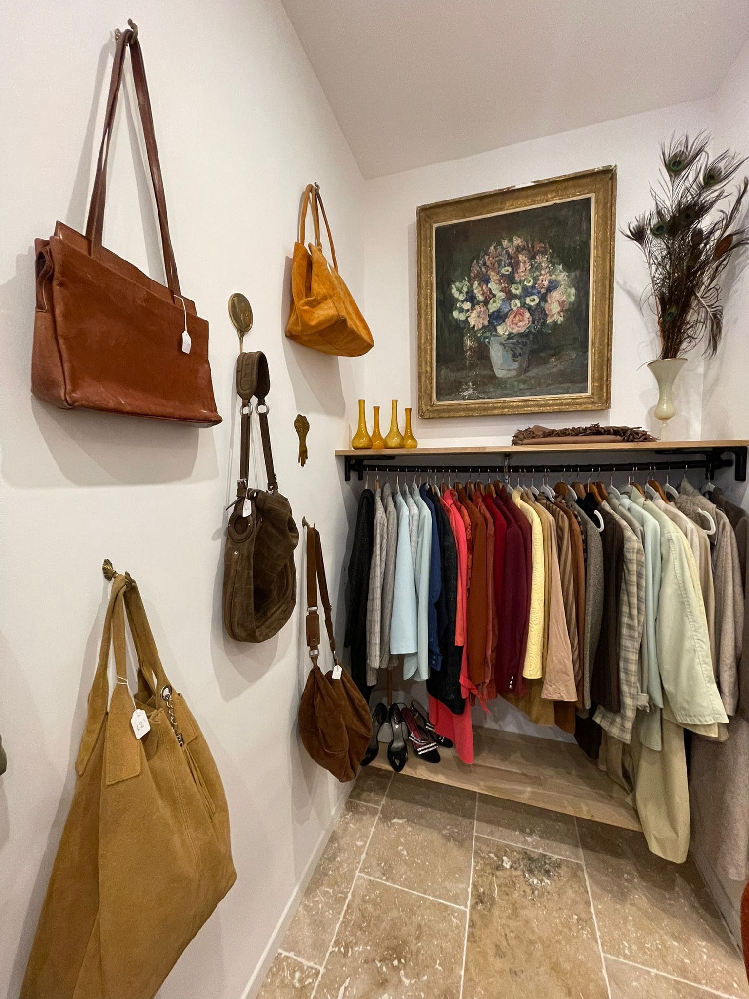 Ein kleines, gemütliches Zimmer, das der Damenmode gewidmet ist. Links an der Wand hängen Taschen aus Leder und Veloursleder. An der linken Wand befindet sich eine Sammlung von Jacken, Mänteln und Hemden in nüchternen und warmen Tönen: Beige, Rot, Blau, Blau, Grün. Darunter befinden sich Schuhe mit hohen Absätzen. An der Wand zeigt ein großes gerahmtes Gemälde einen Blumenstrauß in einer Vase neben einer Vase mit Pfauenfedern und kleinen gelben Karaffen. Die Atmosphäre erinnert an ein schickes und stilvolles Second-Hand-Laden. 