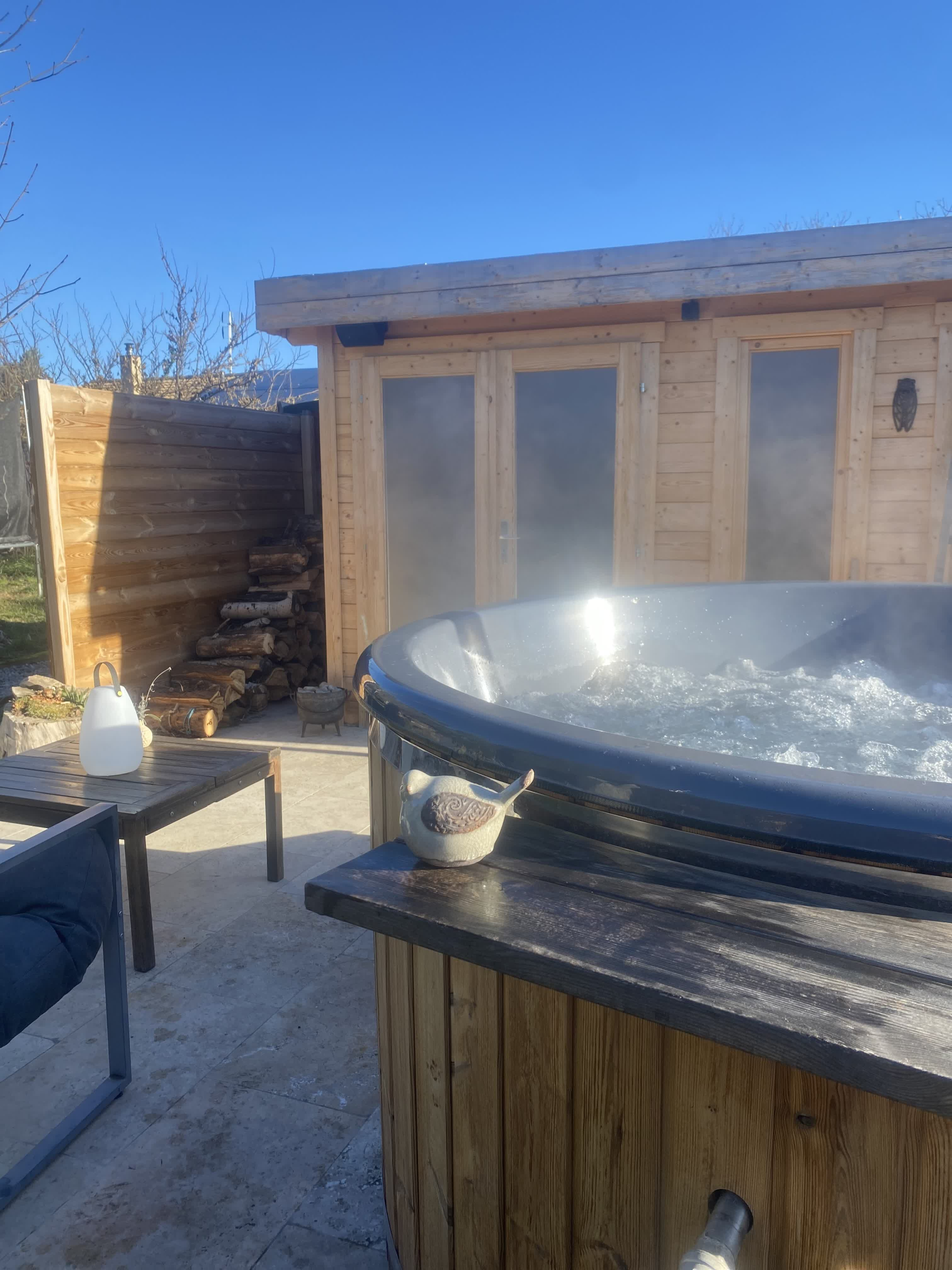 Nordisches Holzbad im Freien, beheizt und rauchend, auf einer sonnigen Terrasse vor einem Holzchalet installiert.