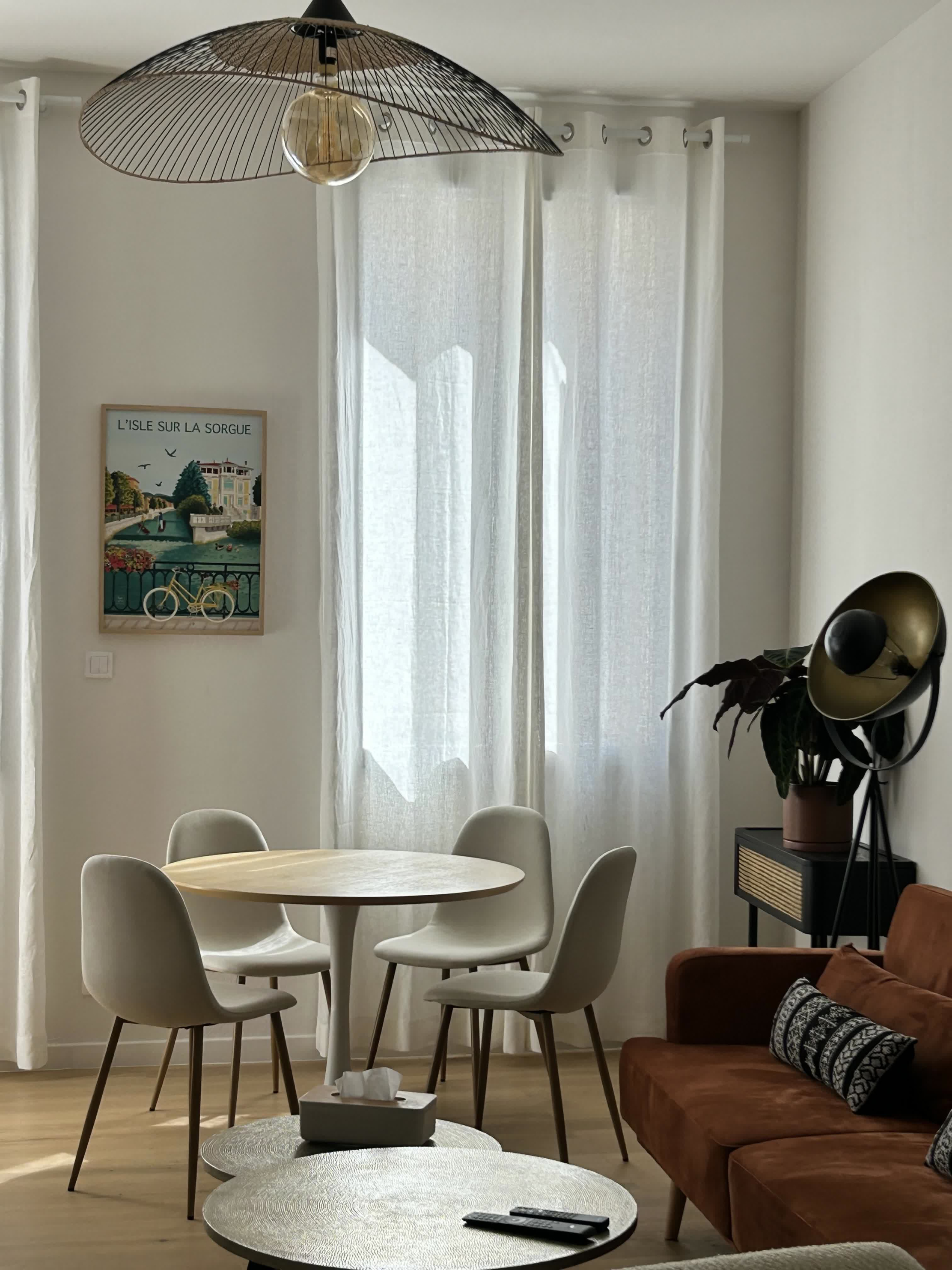 Helles Wohnzimmer mit einem Essbereich, bestehend aus einem runden Tisch und hellen Stühlen, einem braunen Sofa, einem großen verhüllten Fenster und modernem Dekor in Naturtönen.