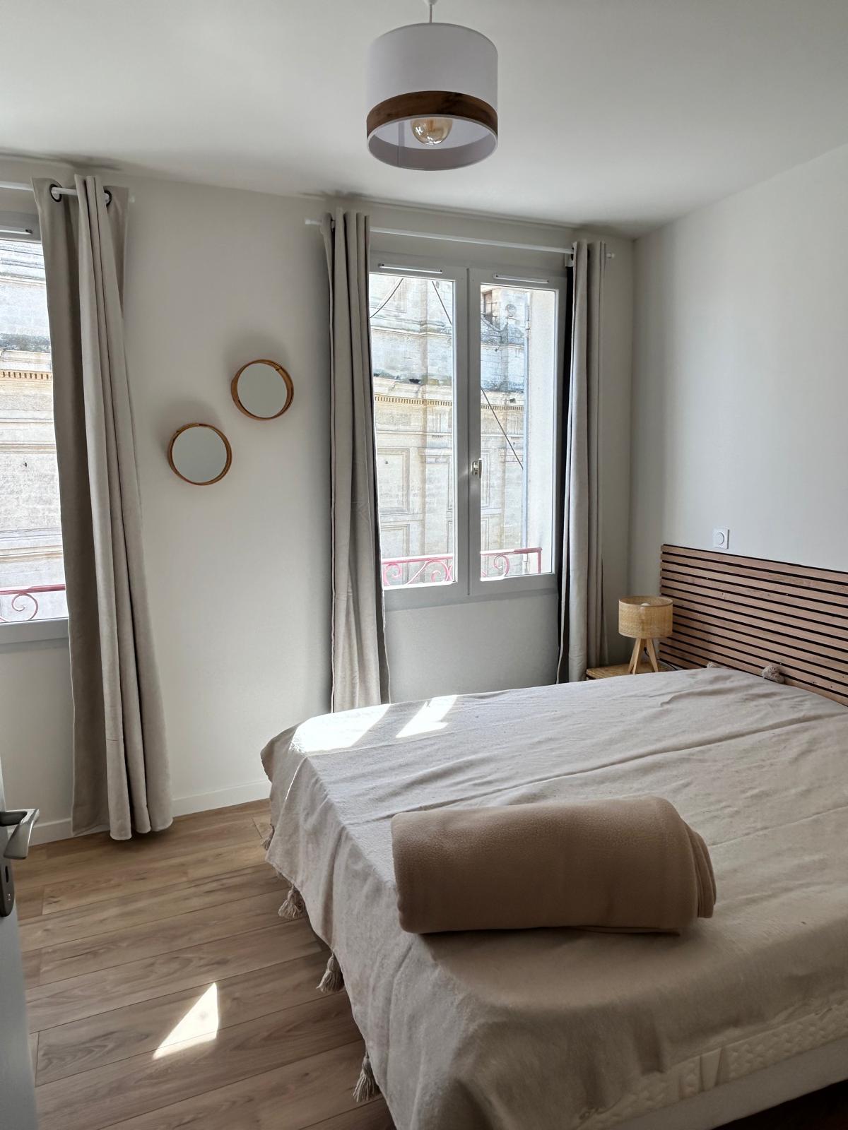 Elegantes und helles Schlafzimmer mit einem Doppelbett aus beigen Textilien, einem Kopfteil aus Holz, Lichtvorhängen und einem großen Fenster, das Tageslicht hereinlässt.