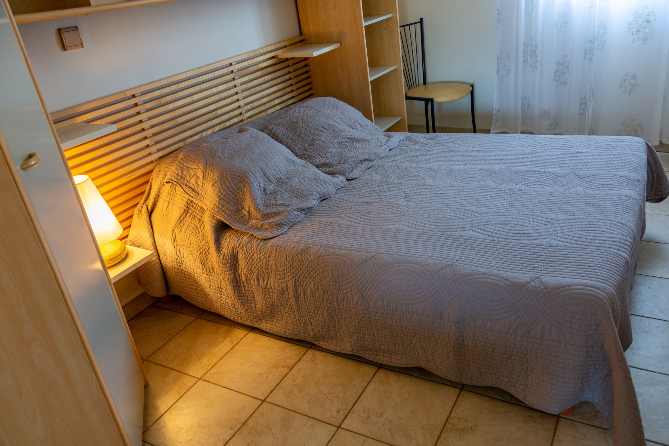 Ruhiges Zimmer mit einem Doppelbett in neutralen Farbtönen, einem hellen Kopfteil aus Holz, eingebautem Stauraum und sanftem Licht durch eine Lampe und ein verdecktes Fenster.
