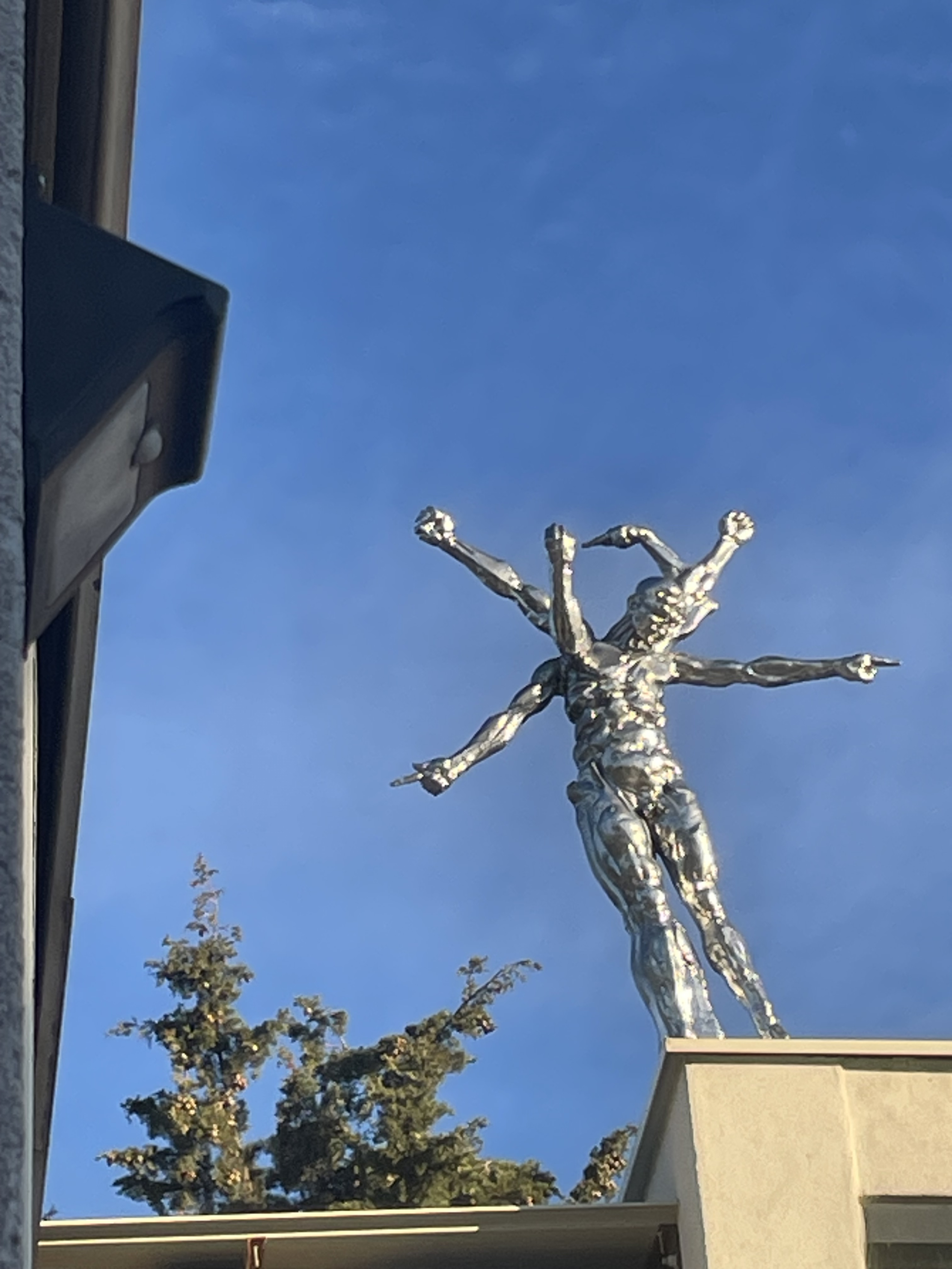 Dieses Foto zeigt eine monumentale Skulptur, die eine menschliche Figur mit mehreren Armen darstellt und in einer glänzenden Metalloberfläche gefertigt ist, die das Licht einfängt. Hoch oben auf einem Gebäude installiert,