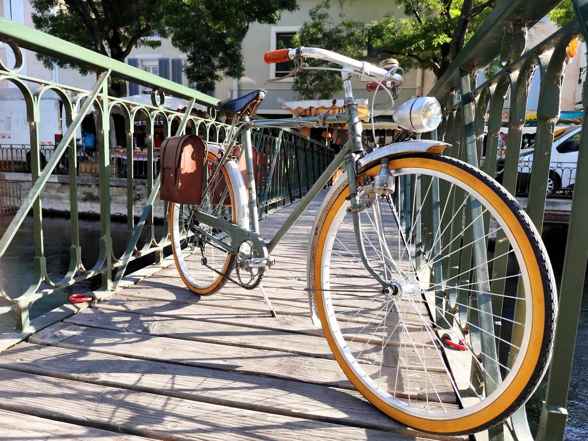Ein Oldtimer-Fahrrad wird auf einer Metallbrücke fotografiert, die die Sorgue überquert. Das Dekor hebt die ikonischen Kanäle der Stadt hervor und lädt Sie ein, sie mit dem Fahrrad zu erkunden.