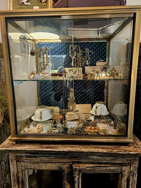 Ein antikes Fenster vereint zahlreichen Modeschmuck: Halsketten, Armbänder, Broschen, Ohrringe und andere Kreationen. Die patinierten Möbel und die sorgfältige Präsentation verstärken den Vintage-Charakter des Ladens.