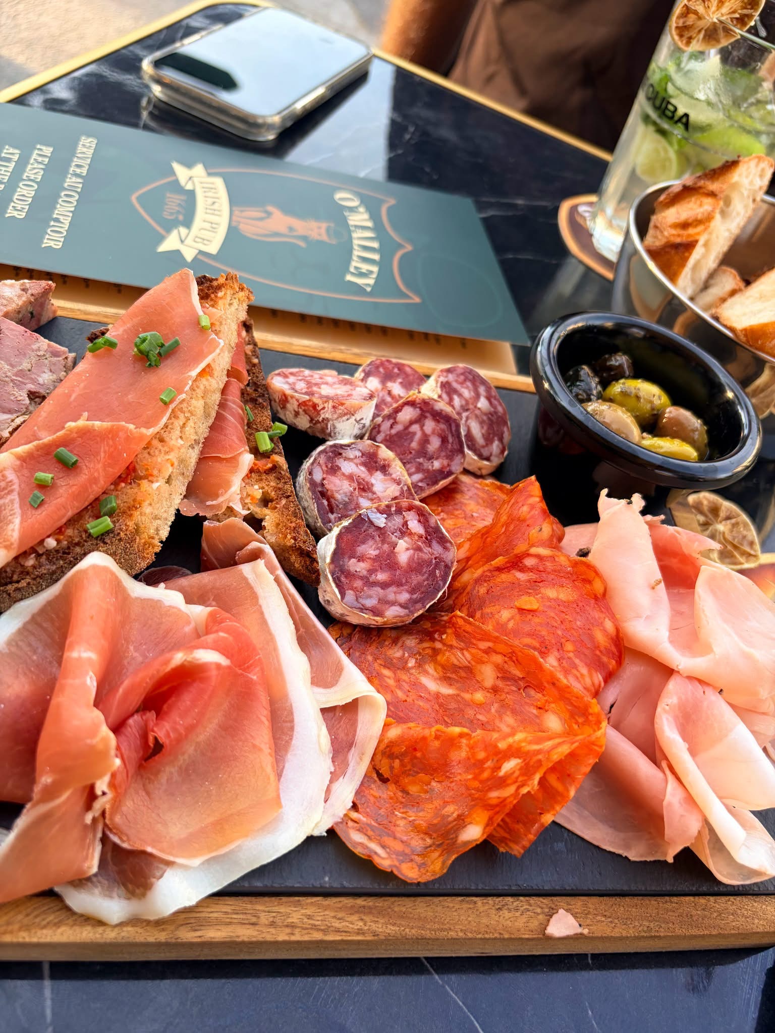 Ein großzügiges Wurstbrett zum Teilen, bestehend aus Schinken, Würstchen und anderen Spezialitäten, begleitet von Oliven und Brot. Im Hintergrund runden ein Cocktail und das O
