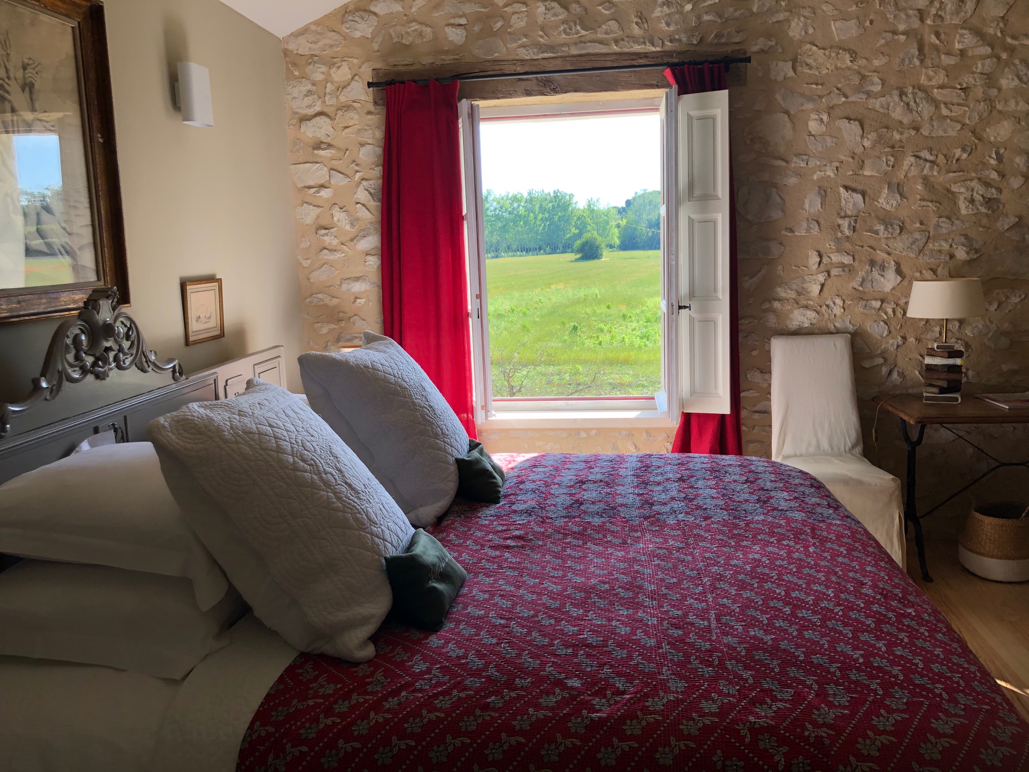 Charmantes Zimmer im Mas de la Sorgue mit unverputzten Steinwänden, roter Bettwäsche und einem großen Fenster mit Blick auf die grüne Landschaft.