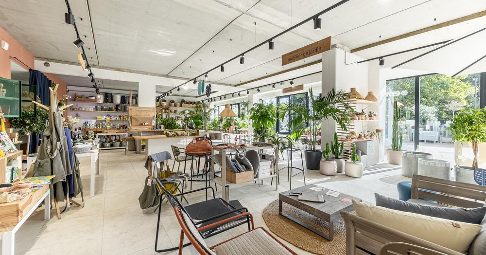 Ein heller und warmer Raum, der grüne Pflanzen, Töpfe, Dekorationsgegenstände und Gartenmöbel kombiniert. Die großen Fenster lassen natürliches Licht herein und unterstreichen die verschiedenen Atmosphären, die im Geschäft herrschen.