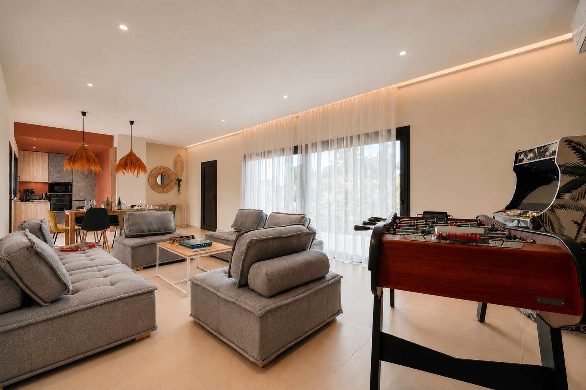 Großes helles Wohnzimmer mit offener Küche, Essbereich und freundlicher Lounge. Das Zimmer hat auch einen Tischfußball und einen Spielautomaten, ideal, um Momente mit Familie oder Freunden zu teilen.