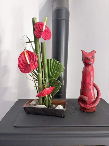 Blumenarrangement im Ikebana-Stil, das rote Anthurien, Bambusstängel und grafisches Laub kombiniert und in einem rechteckigen Behälter mit Kieselsteinen angeordnet ist.